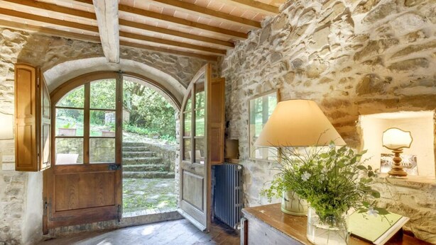 Interior rústico de pedra com uma porta arqueada que dá para um jardim, uma mesa de madeira com uma lâmpada e um vaso de flores, e um radiador.