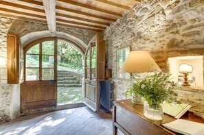 A rustic stone interior with an arched doorway leading to a garden, a wooden table with a lamp and a vase of flowers, and a radiator. - Wine & History in Montalcino- Stay in an ancient mill (Castelnuovo dell'Abate)
