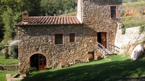 Um edifício de pedra com telhado em telhas, porta de madeira e pequena escadaria que dá acesso a um segundo andar.