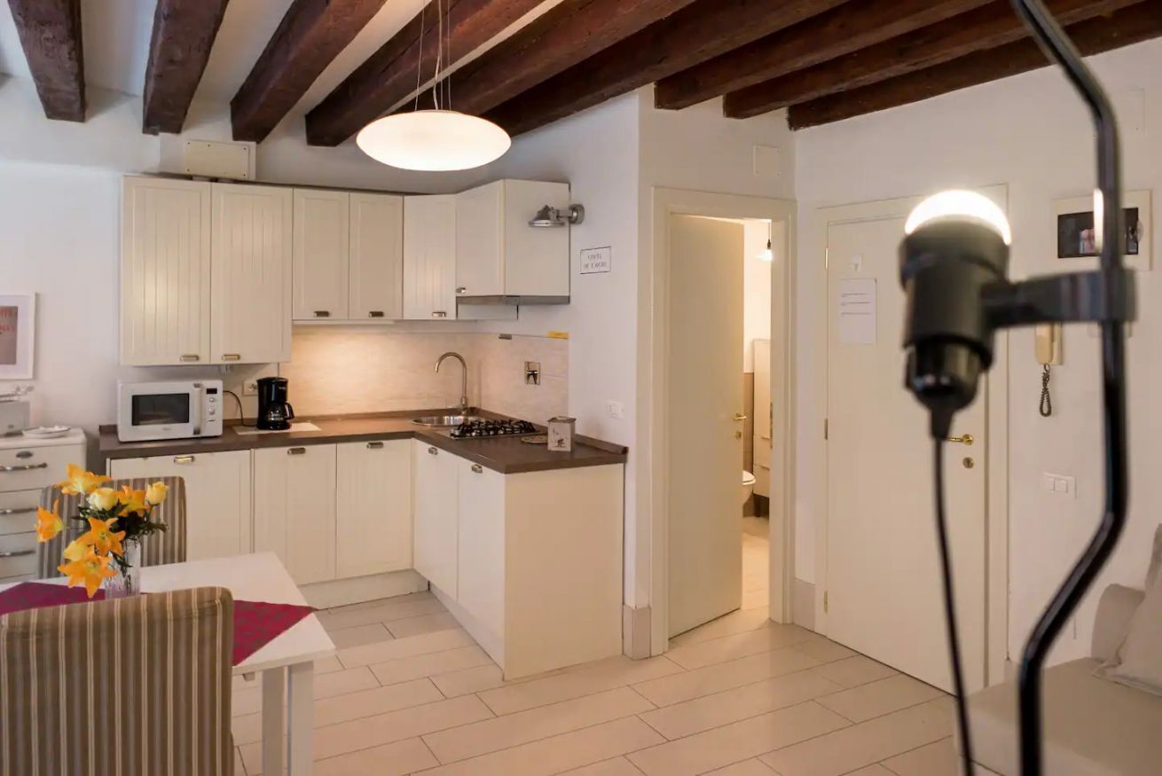 A compact kitchen with white cabinets, a microwave, a coffee maker, and a sink. There is a dining area with a table and chairs, and a vase with flowers.
