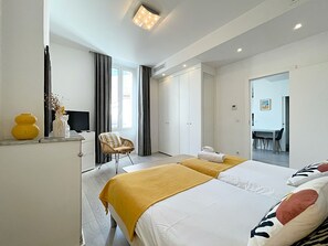 A modern hotel room with two beds, a TV, a chair, and a dining area visible through an open door. - Lovely and Modern 1 Bedroom City Centre Cannes (Cannes)