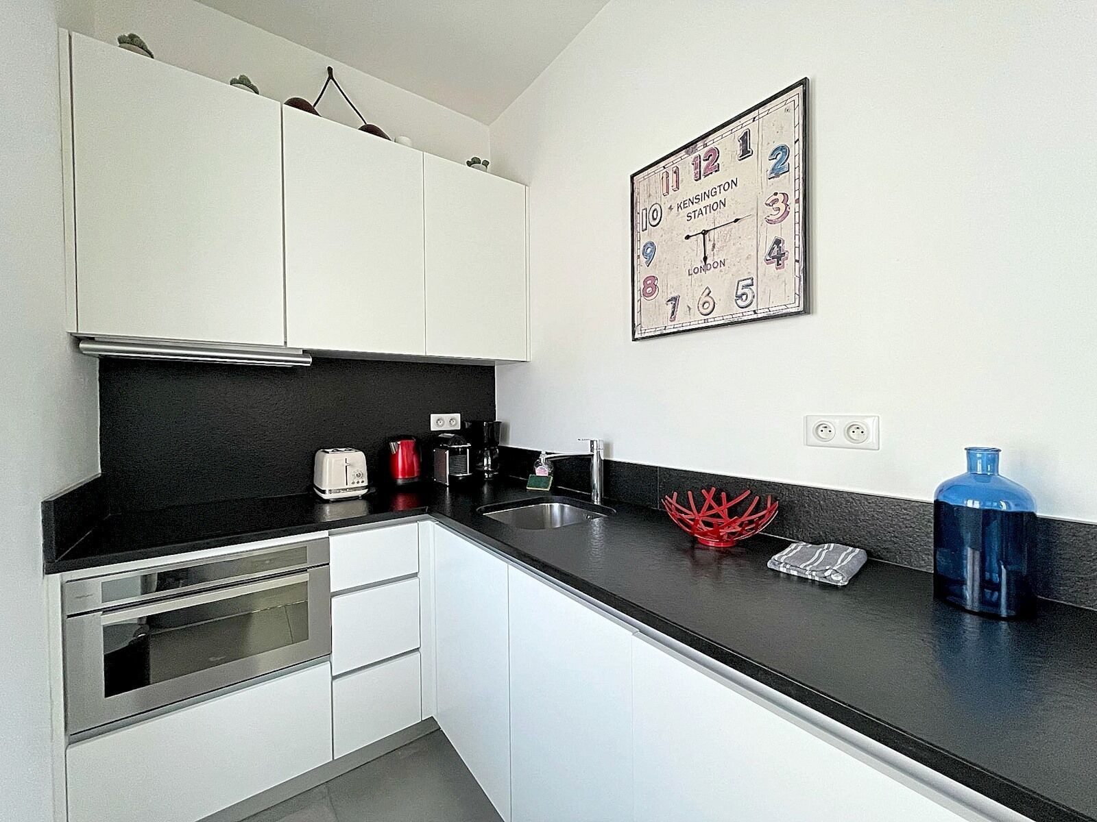 A modern kitchen with white cabinets, a black countertop, a coffee maker, a toaster, and a red decorative bowl.