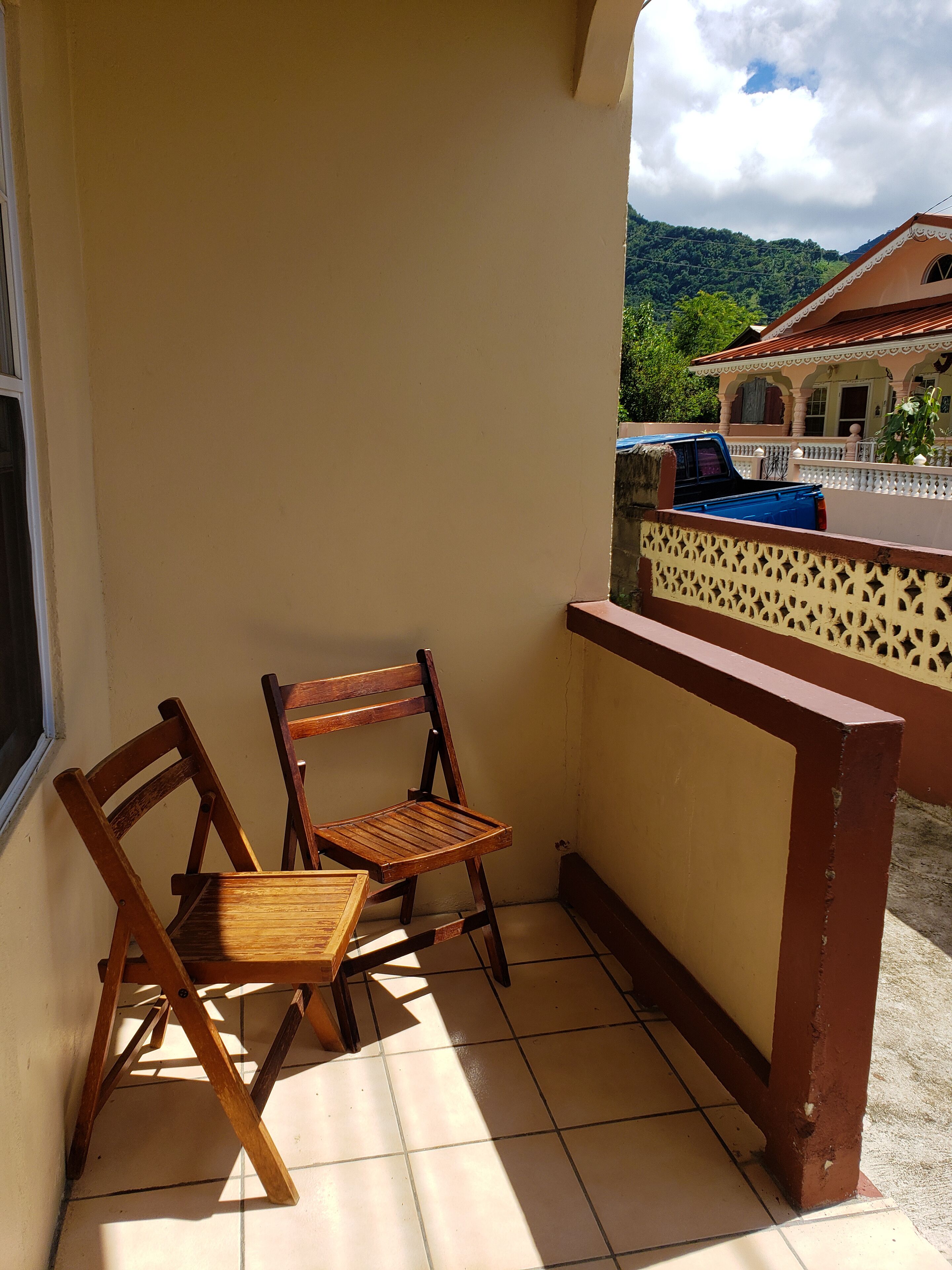 A balcony with two wooden chairs, a tiled floor, and a view of a house and mountains in the background.