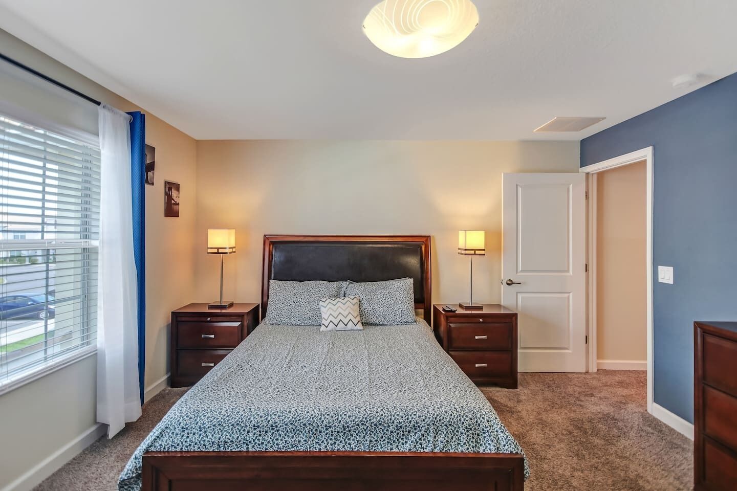 A bedroom with a bed, two bedside tables, a nightstand, a window with blinds, and a door.