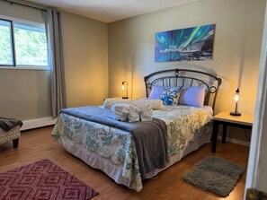 A neatly made bed with a patterned bedspread, two bedside lamps, a window with curtains, and a painting of the aurora borealis on the wall. - Granite Getaway - Quiet, Comfortable Place Offering 10% OFF ON TOURS! (Juneau)