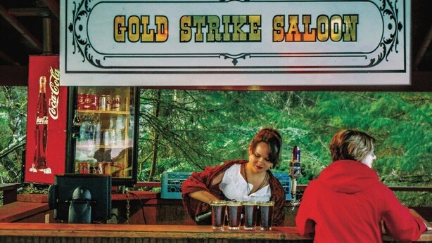 Eine alte Coca-Cola-Automat und ein Schild mit der Aufschrift 'Gold Strike Saloon' in ländlicher Umgebung.