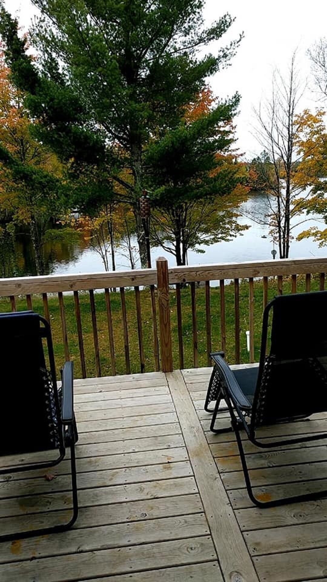 Une terrasse en bois avec deux chaises noires, donnant sur une étendue d’eau, le tout entouré d’arbres aux feuilles d’automne.