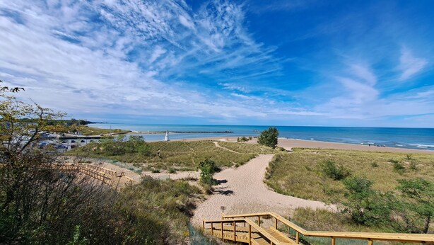 Ein Strand mit einer hölzernen Prominade, die zum Ufer führt, umgeben von grasbewachsenen Dünen und spärlicher Vegetation.