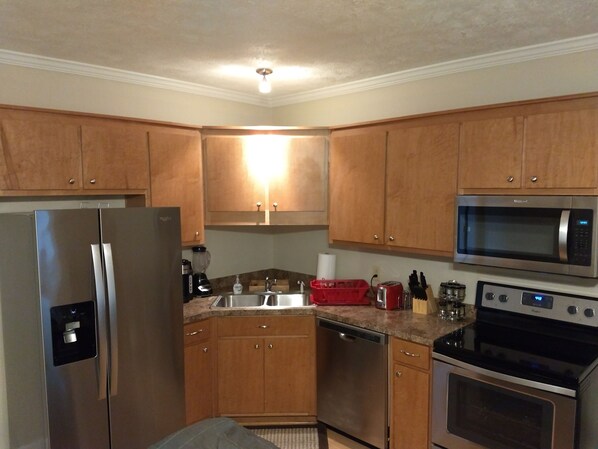 A modern kitchen with wooden cabinets, stainless steel appliances including a refrigerator, microwave, and oven, and a granite countertop.