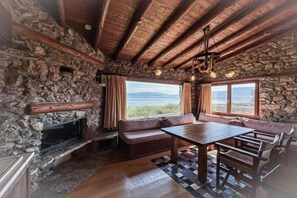 Een rustiek interieur met stenen muren, een open haard, een houten plafond en een groot raam met uitzicht op het landschap.