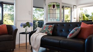 A living room with a black leather sofa, a small side table with a vase of flowers, and a view of the outdoors through large windows. - Aggies Way - dog friendly with  panoramic views (Daylesford)