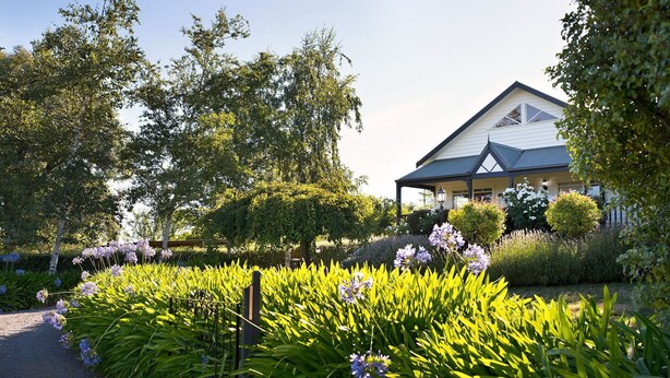 A house with a covered porch, surrounded by lush greenery and flowering plants. - Aggies Way - dog friendly with  panoramic views (Daylesford)