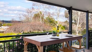 A wooden table with two wine glasses and a bottle, set on a balcony with a view of trees and houses. - Aggies Way - dog friendly with  panoramic views (Daylesford)