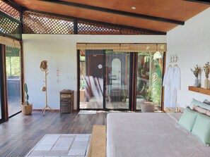Une chambre moderne dotée d’un grand lit, d’un plafond en bois, de portes-fenêtres coulissantes et offrant une vue sur la verdure environnante.