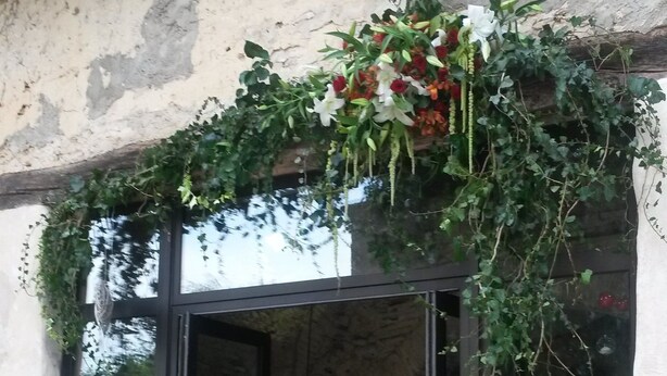 A glass door entrance to a room with a table and chairs inside, a hanging plant above the door, and a brick wall in the background.