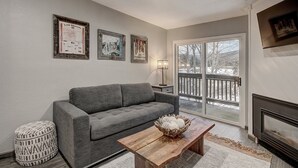 A living room with a gray sofa, a wooden coffee table with decorative items, a patterned ottoman, and a fireplace. There are three framed pictures on the wall and a large window with a view of a snowy deck. - 2bd_mw_315 (Frisco)