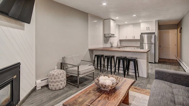 A modern kitchen with a central island, bar stools, and a dining area with a wooden table and a woven ottoman. - 2bd_mw_315 (Frisco)