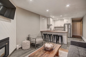 A modern kitchen with a central island, bar stools, and a dining area with a wooden table and a woven ottoman.