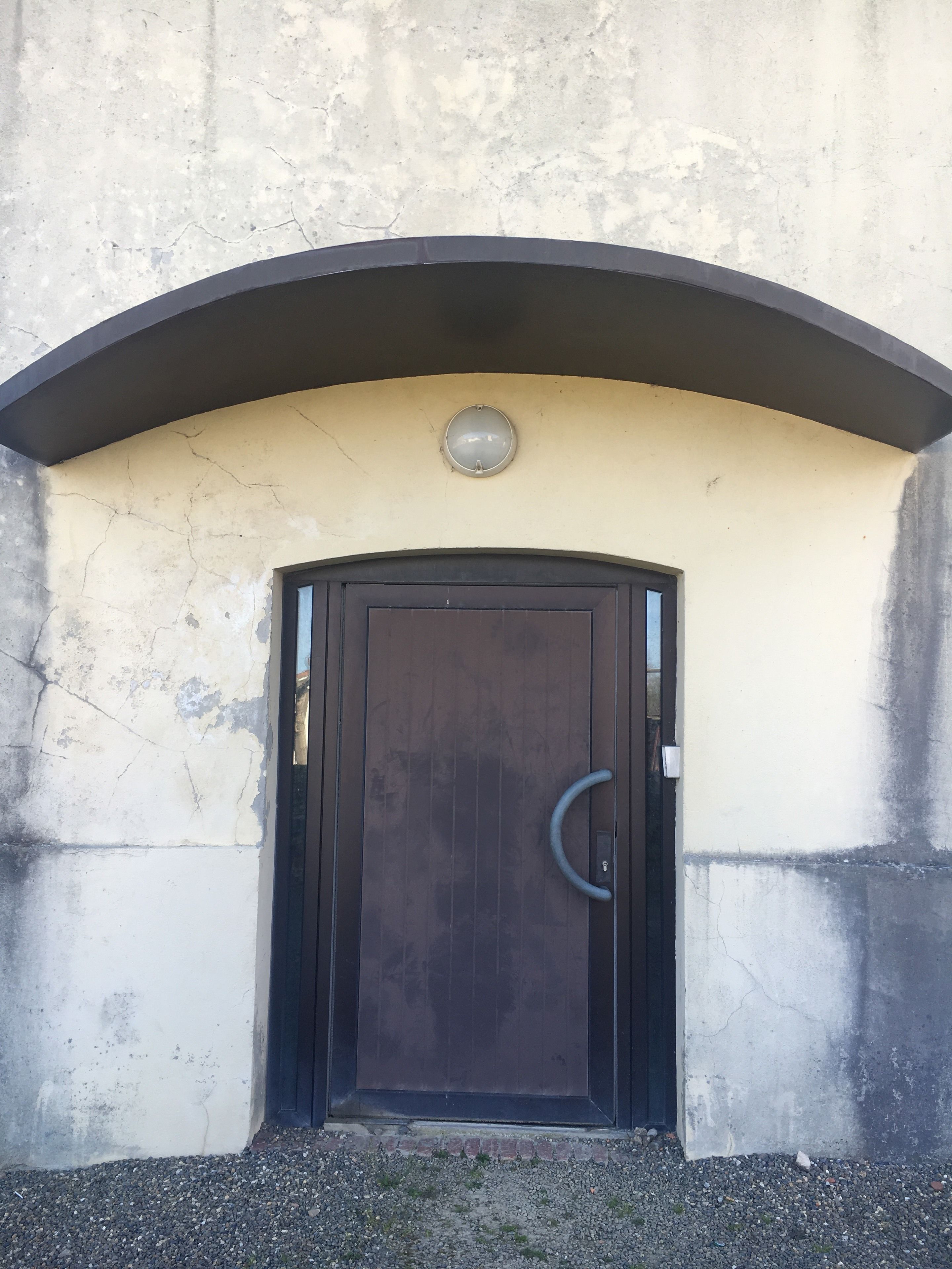 Une porte dotée d’une poignée circulaire, un luminaire rond au-dessus, et un mur en béton présentant une fissure.
