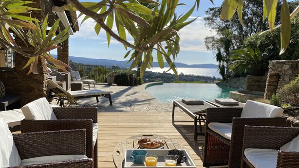A wooden deck with wicker furniture, a breakfast tray, and a view of a swimming pool and mountains. - Very nice villa with infinity pool, sea view, harbor, Cavalaire (Cavalaire-Sur-Mer)