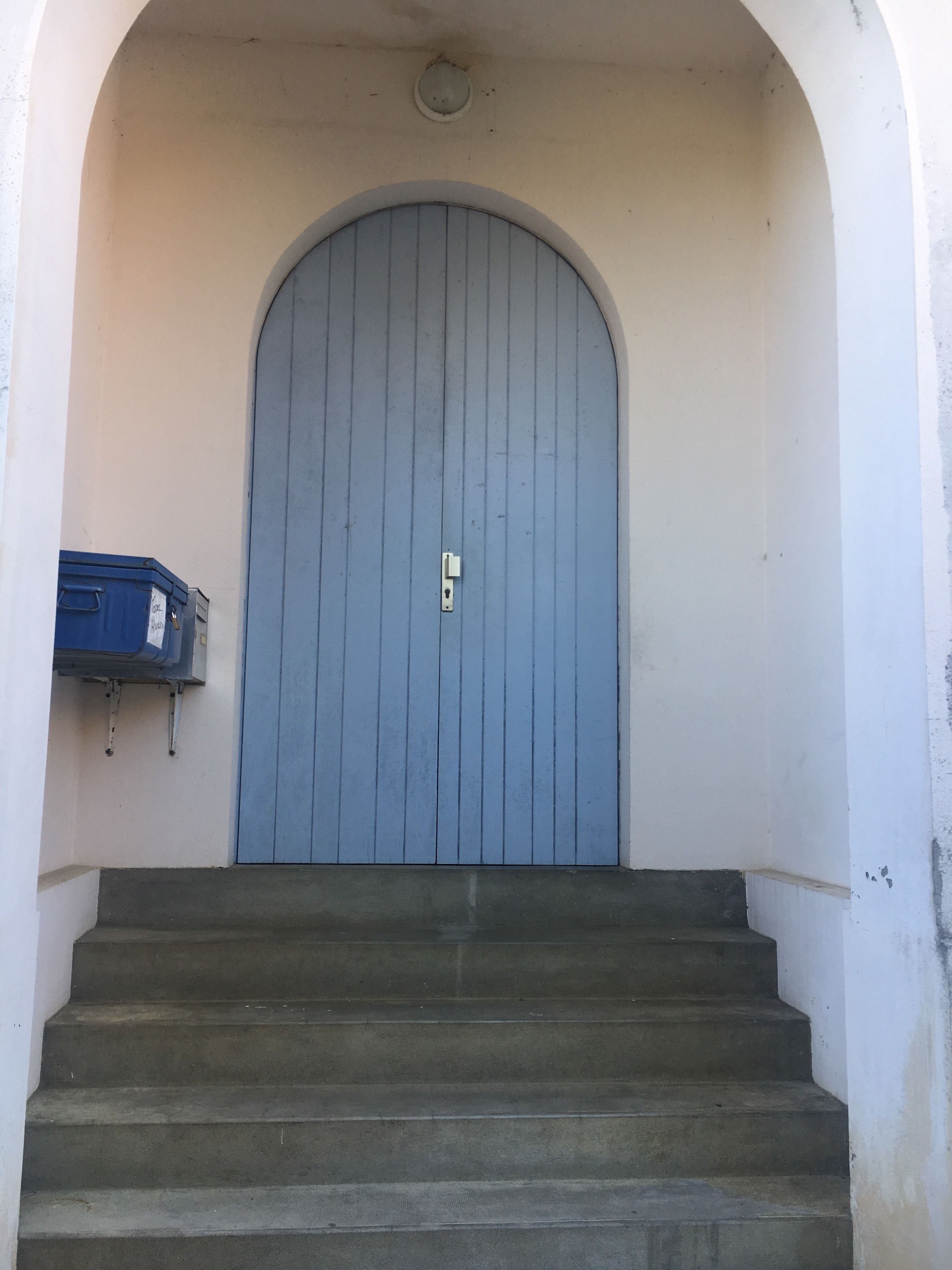 Una puerta azul con manija de metal, entre paredes blancas y una escalera de concreto que sube hacia ella.
