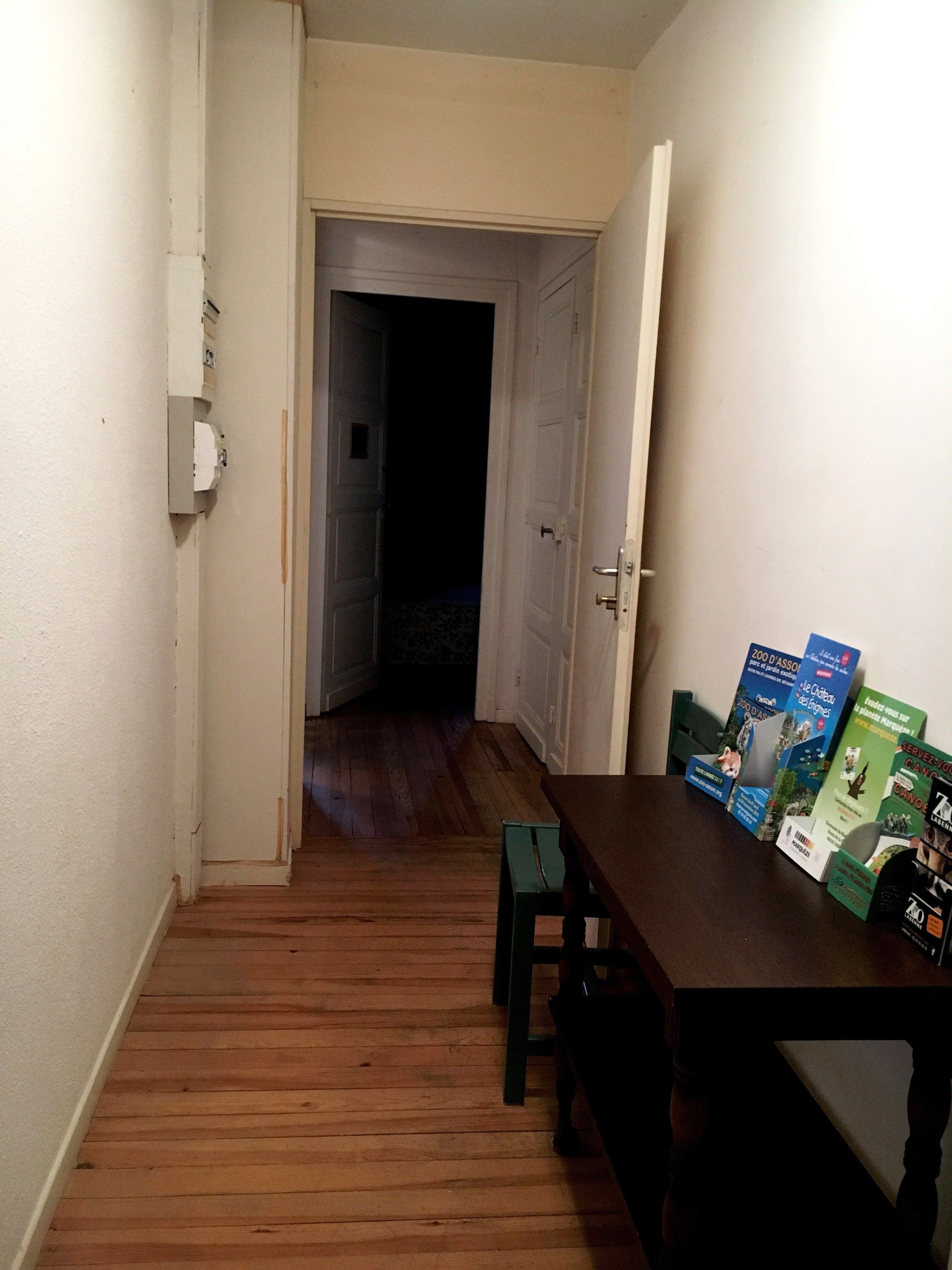 Un couloir étroit, doté d’un plancher en bois, mène à une pièce sombre. Une table sur laquelle sont disposées des brochures est placée contre le mur.