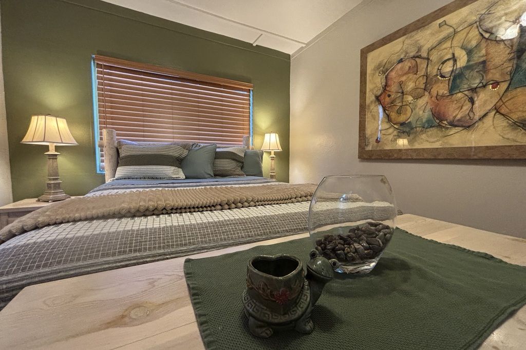 Un dormitorio con una cama, dos lámparas de noche, un ventanal con persianas, un cuadro en la pared y un cuenco decorativo sobre un tapete verde.