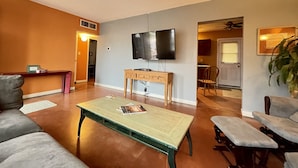 Sala de estar con televisor, una mesa de centro y un sofá. Hay una puerta que da a un comedor.