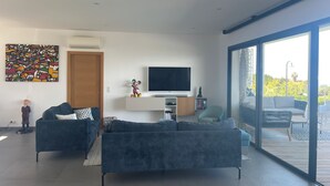 A modern living room with a large TV, two sofas, a bookshelf, and a sliding glass door leading to an outdoor area. - Contemporary, single-story villa with an exceptional view of the Gulf of Ajaccio, (Albitreccia)