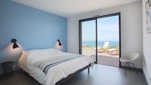 A bedroom with a bed, two wall-mounted lights, a chair, and a large sliding glass door leading to an outdoor area with a view of the sea. - Contemporary, single-story villa with an exceptional view of the Gulf of Ajaccio, (Albitreccia)