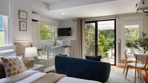 A modern hotel room with a bed, a sofa, a dining area with a table and chairs, a kitchen area, and large windows offering a view of greenery.