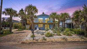 A two-story house with a balcony, surrounded by palm trees and a gravel driveway.