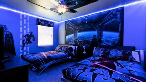 Quarto temático de Star Wars, com uma cama, duas mesas de cabeceira, um ventilador de teto e uma janela com persianas.