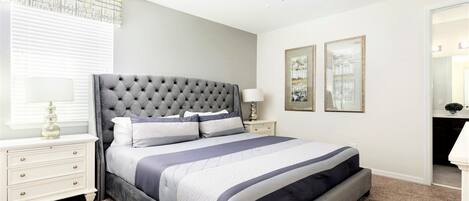 Une chambre à coucher avec une tête de lit capitonnée, un lit recouvert d’une couvre-lit à rayures, une table de chevet avec une lampe, une fenêtre avec des stores et deux tableaux encadrés accrochés au mur.