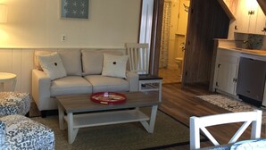 Una sala de estar acogedora con un sofá, una mesa de centro y otomanas estampadas. Se ve una mesa de comedor con un camino de mesa azul estampado. El piso es de madera y tiene un ventilador de techo.