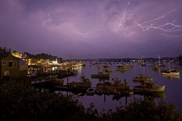 Un port de plaisance de nuit, avec des bateaux amarrés, un bâtiment illuminé et des éclairs dans le ciel.