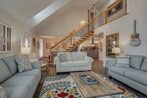 A spacious living room with a high ceiling, a staircase leading to an upper level, and a kitchen in the background.