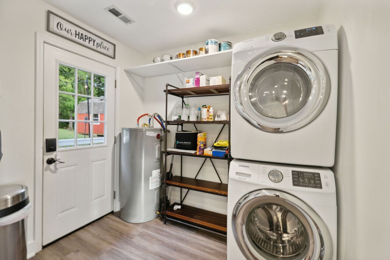 Un cuarto de lavandería con lavadora y secadora de carga frontal, repisas con productos de limpieza, un bote de basura y una puerta que da a la vista exterior.