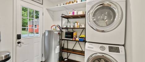 Un cuarto de lavandería con lavadora y secadora de carga frontal, repisas con productos de limpieza, un bote de basura y una puerta que da a la vista exterior.