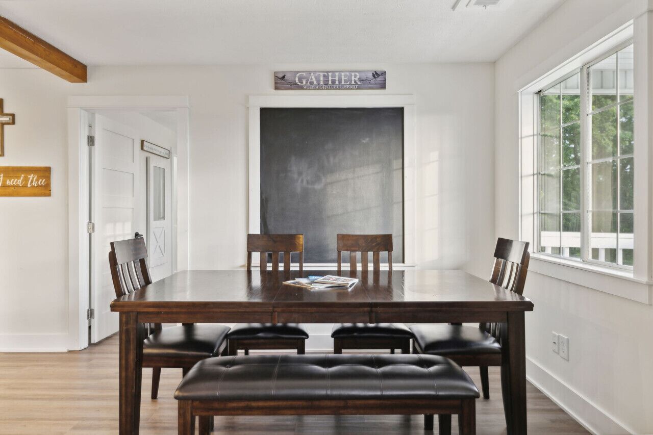 A dining room with a large wooden table, four chairs, and a bench. There is a sign above the table that reads "GATHER."