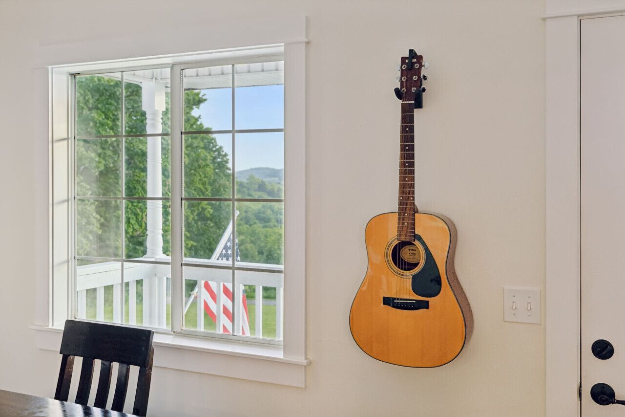 Una habitación con ventana que da a una terraza, una bandera estadounidense, una guitarra de madera colgada en la pared y una silla cerca de la ventana.