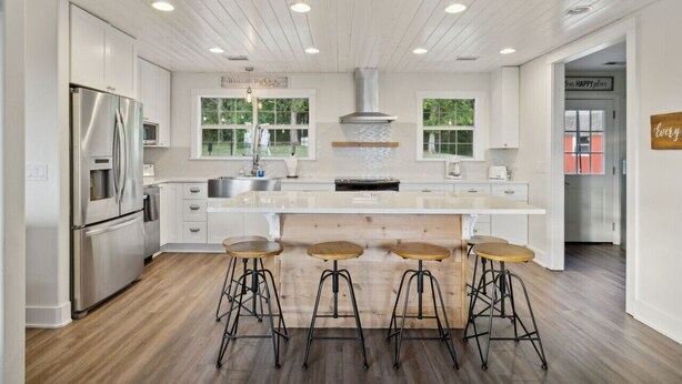 A modern kitchen with white cabinetry, stainless steel appliances, a central island with stools, and a view of the outdoors through large windows.
