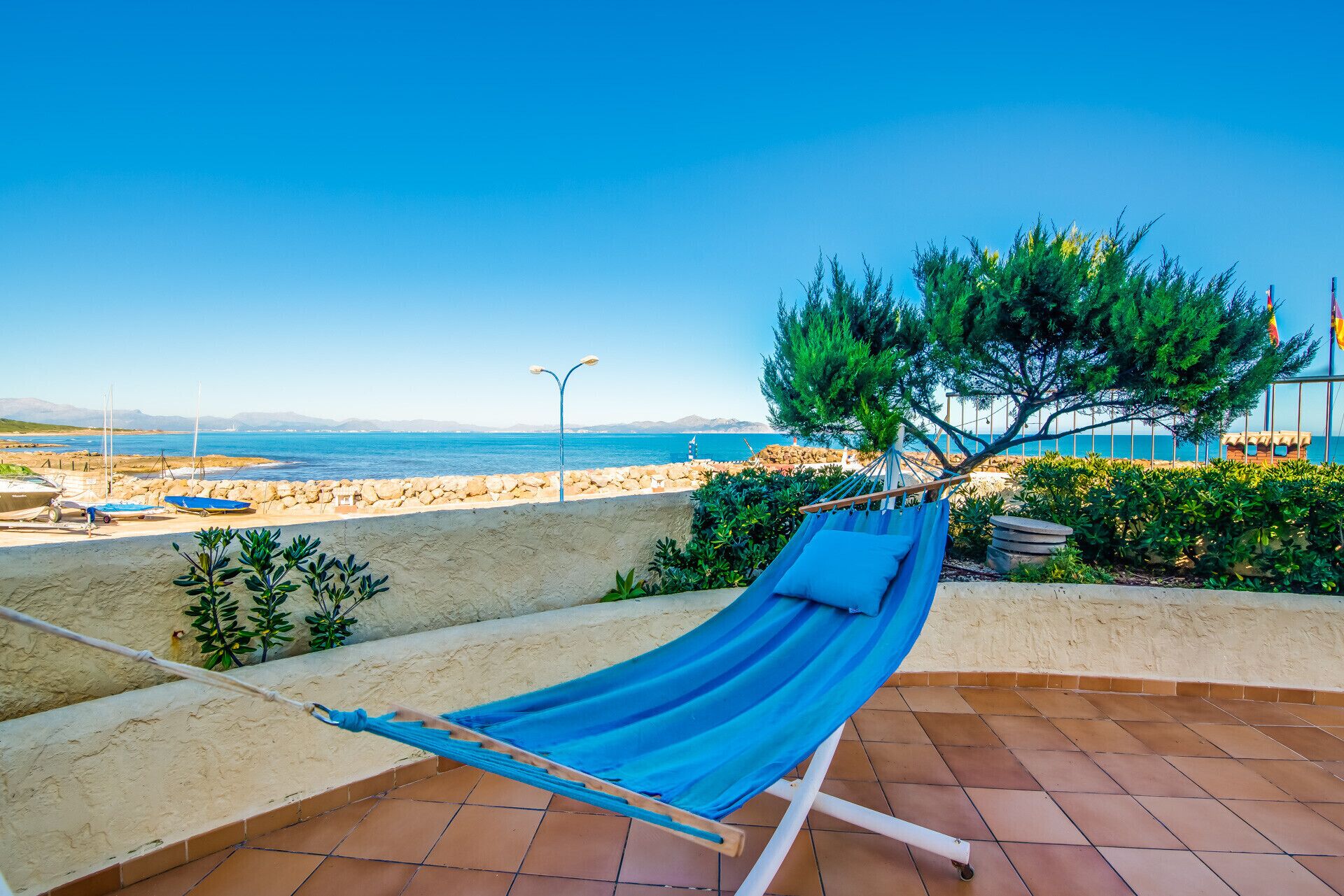 A hammock on a tiled patio overlooking a beach with boats and a clear sky.