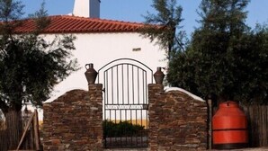 Une piscine dans laquelle se reflète un bâtiment à la façade blanche, au toit recouvert de tuiles rouges, aux murs en pierre et à l’entrée fermée par un portail.