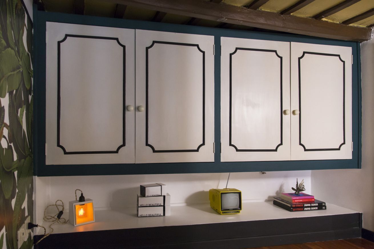A white cabinet with black trim and four doors, a small white shelf with a yellow TV, and a wall-mounted power outlet.