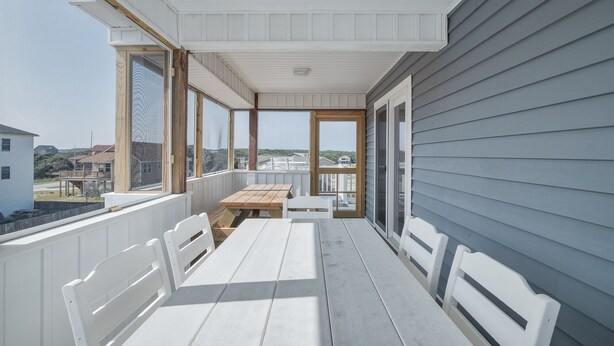 En overdækket veranda med et bord og stole, store vinduer og udsigt over det omkringliggende landskab.