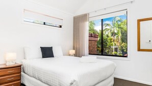 Une chambre d’hôtel avec un grand lit, une table de chevet avec une lampe, une fenêtre donnant sur des palmiers et une tête de lit en bois.
