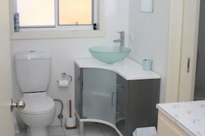 A bathroom with a toilet, a glass basin sink, a toilet paper holder, a broom, and a trash bin.