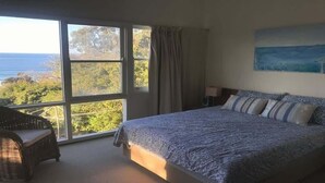 Une chambre avec une grande fenêtre, un lit recouvert d’une literie à motifs, une chaise en osier et un tableau accroché au mur.
