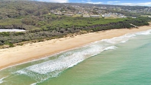 Vue aérienne d’une plage avec un rivage sablonneux, une petite crique et une zone résidentielle avec des maisons en arrière-plan.